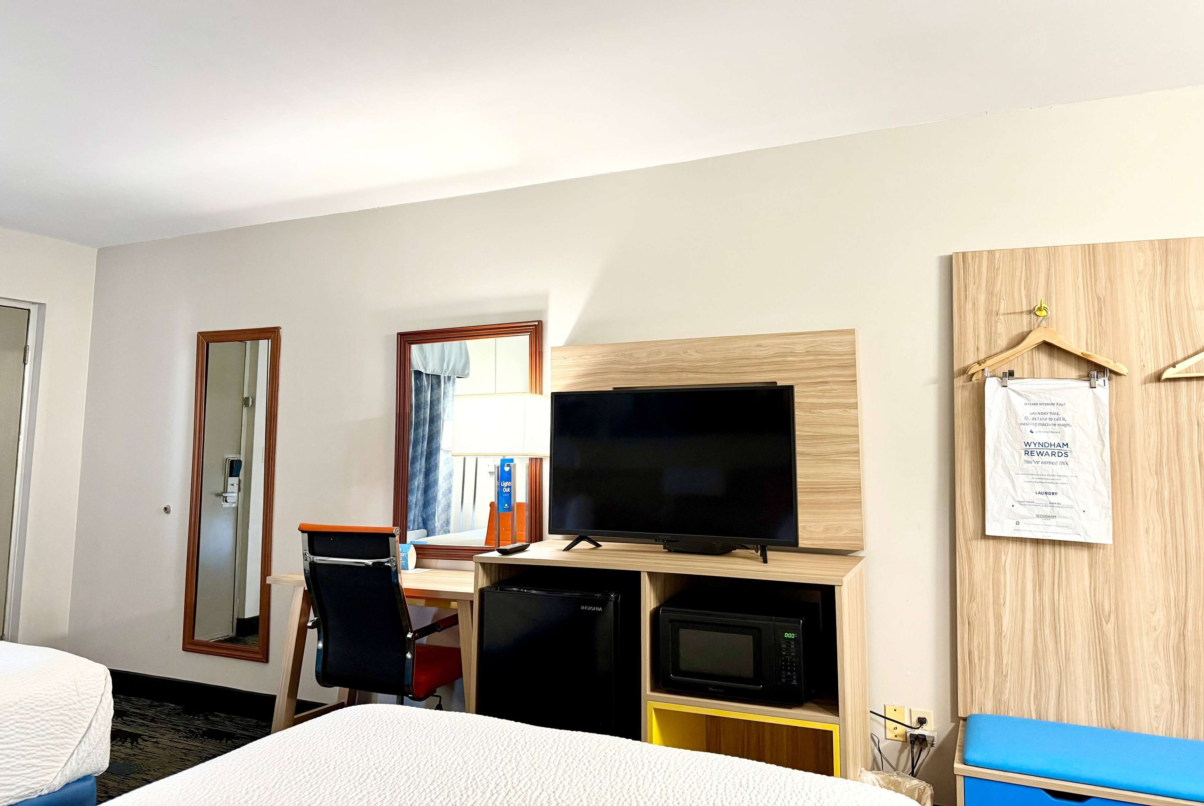 A hotel room with a bed, a desk with a computer and a flat-screen TV, a microwave, a mini-fridge, and a blue storage unit.