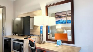 A hotel room with a TV, a desk with a lamp, a microwave, a chair, and a bed with an orange headboard.