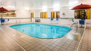 An indoor swimming pool with blue tiles, white walls, and blue lounge chairs. There are red umbrellas and a ceiling light fixture. - Baymont by Wyndham Helena (Helena)