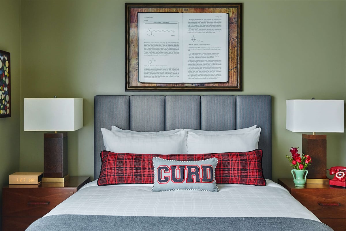 A neatly made bed with a pillow labeled "CURD," a framed document on the wall, and a vintage red telephone on the nightstand.
