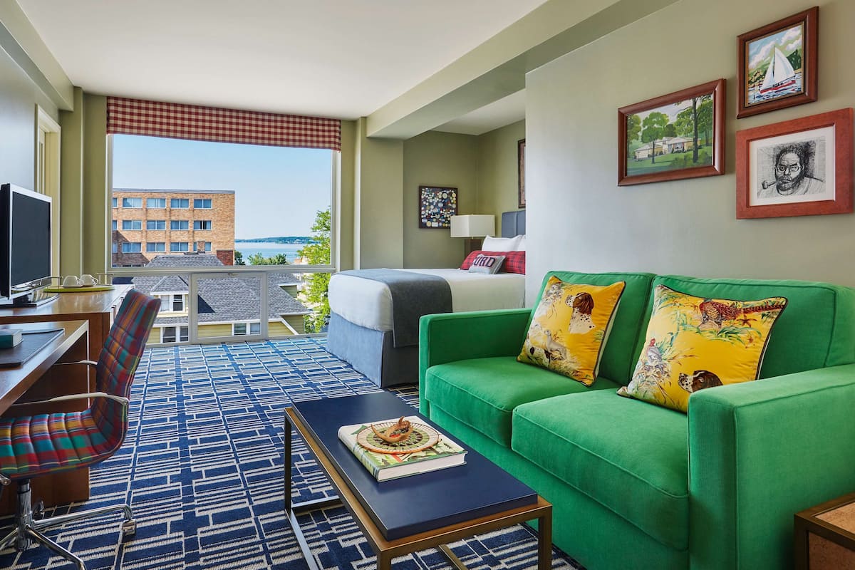 A hotel room with a bed, green sofa, and a desk with a chair. There are framed pictures on the wall and a view of a building outside.