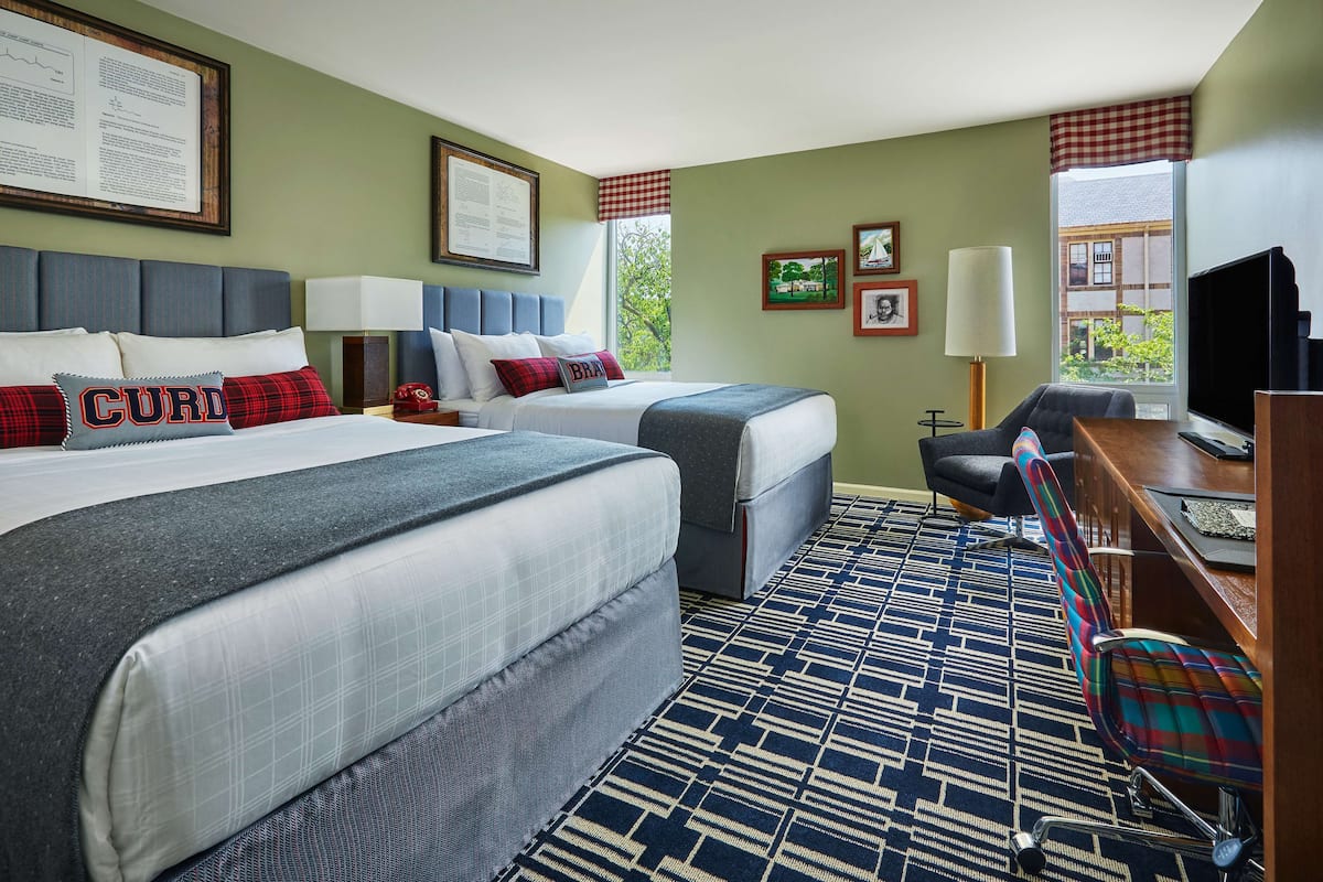 A hotel room with two beds, a desk, a chair, a TV, and a window with a view of greenery.