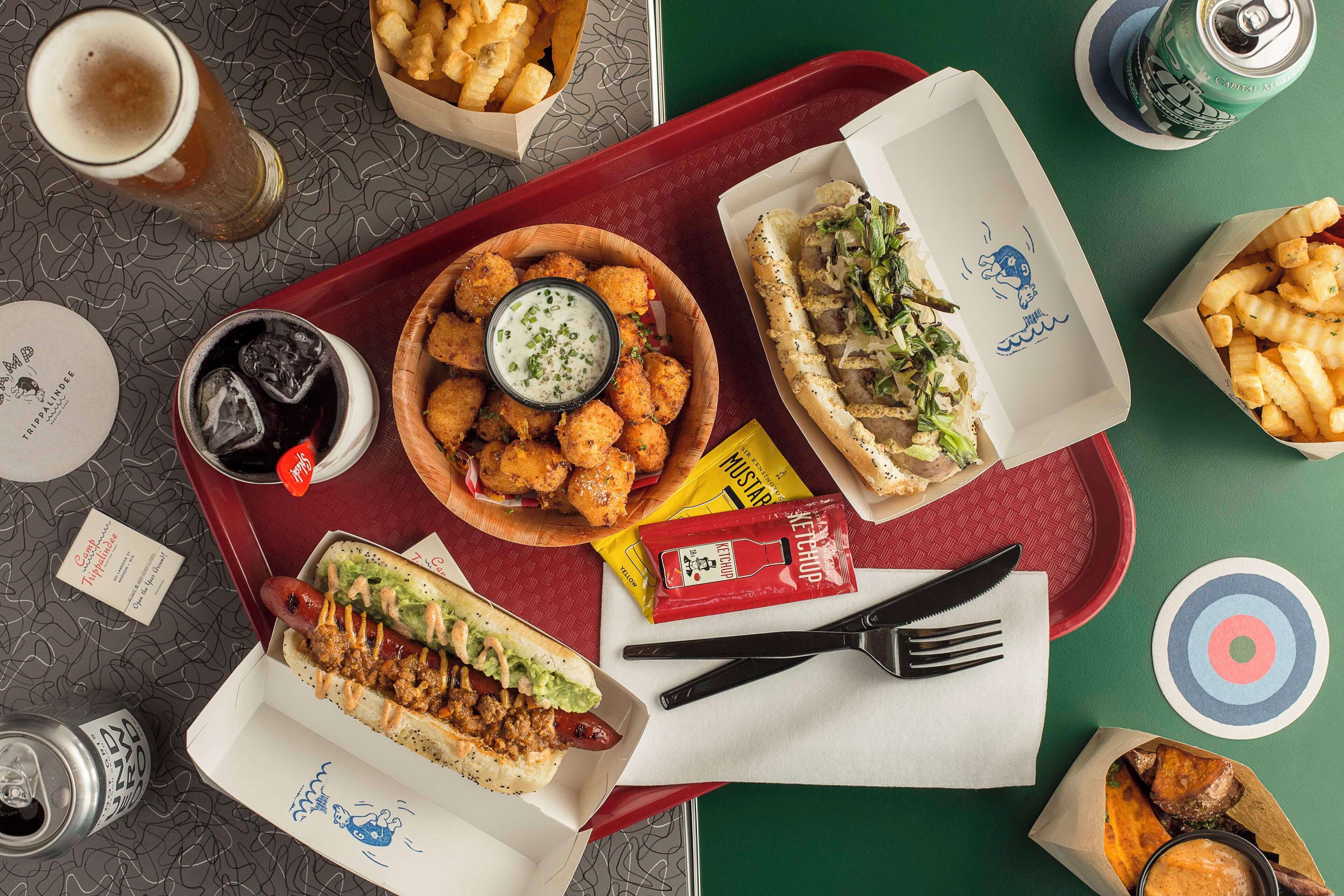 Una bandeja con comida rápida variada, incluyendo un perro caliente, croquetas de papa y un sándwich, junto con salsas y una bebida.