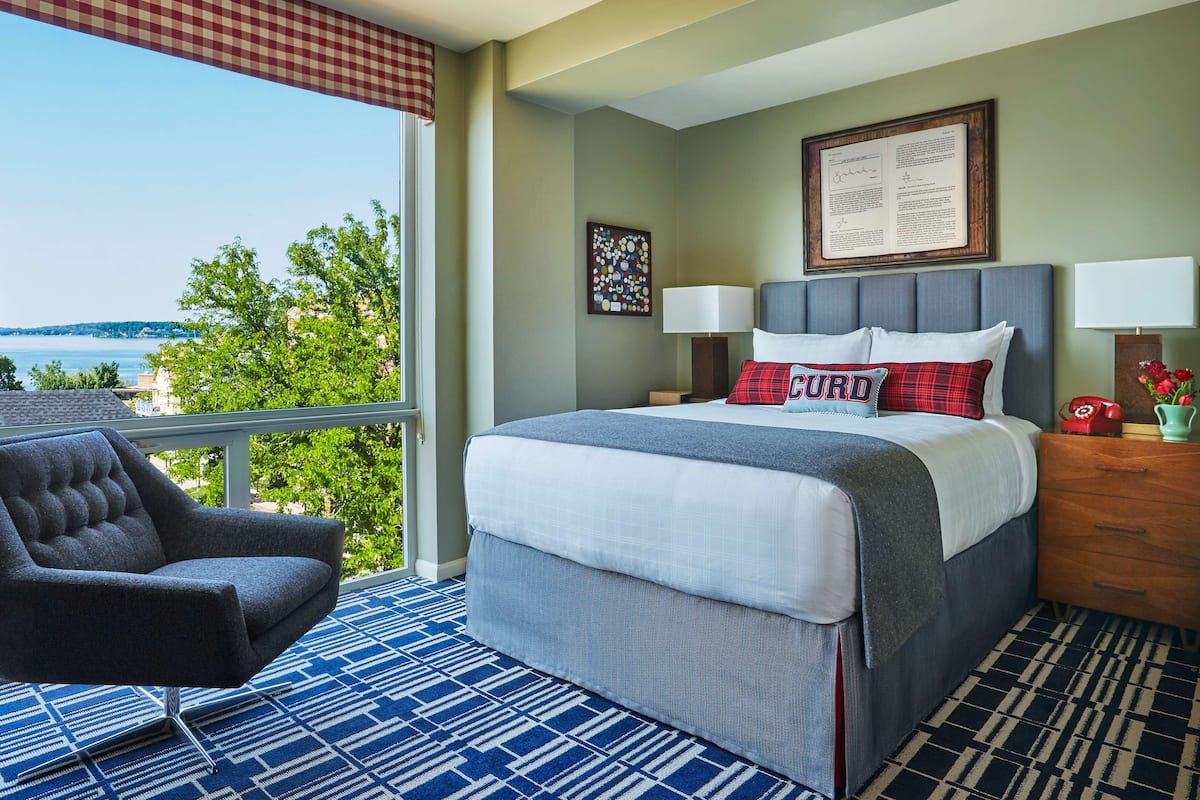 A hotel room with a large bed, two bedside lamps, a nightstand with a red telephone, a chair, and a view of the sea through a large window.