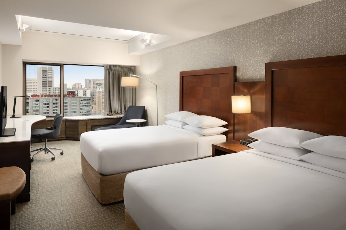 Ein Hotelzimmer mit zwei Betten, einem Schreibtisch, einem Stuhl und einem großen Fenster mit Blick auf die Stadt.
