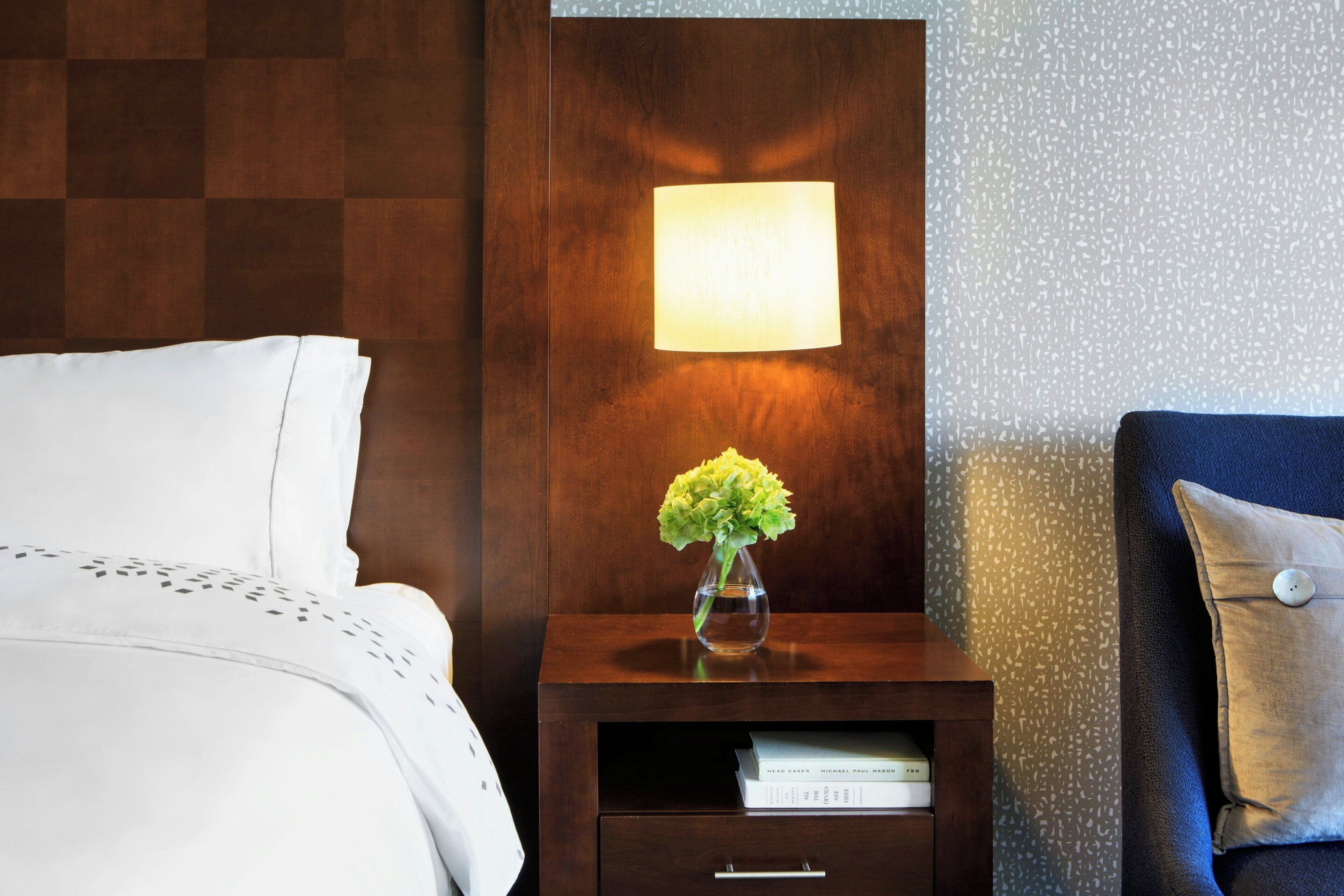 A neatly made bed with white linens, a small wooden nightstand with a lit wall lamp, a vase with flowers, and a blue pillow.