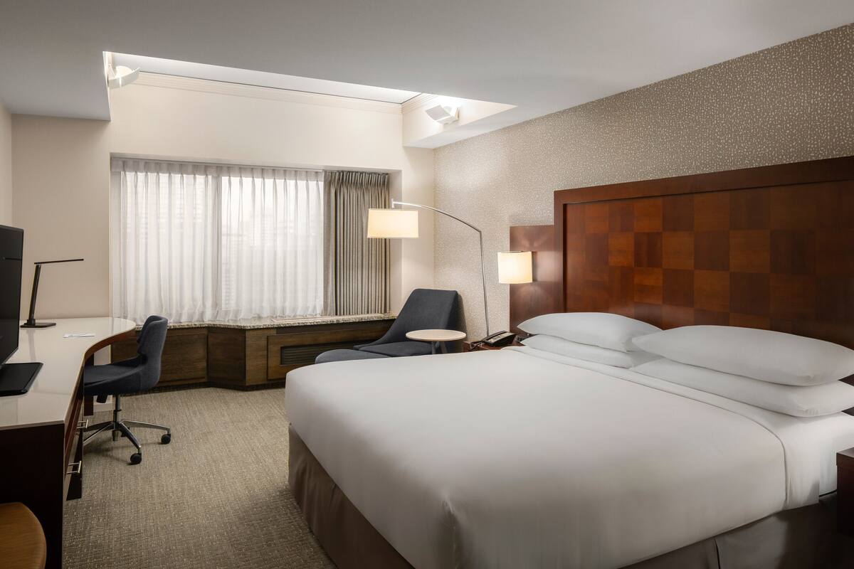 Ein Hotelzimmer mit einem großen Bett, einem Schreibtisch mit Stuhl, einer Lampe und einem Fenster mit Vorhängen.