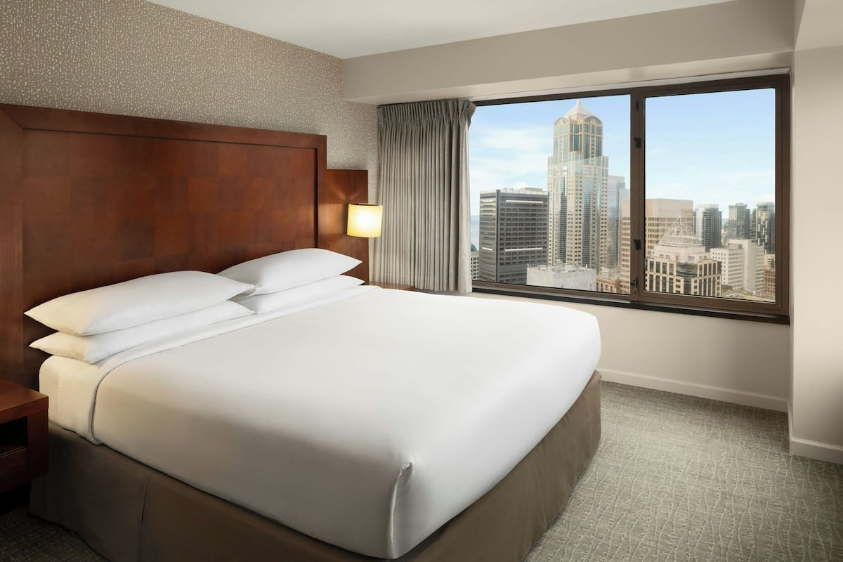 Ein Hotelzimmer mit einem großen Bett, einem hölzernen Kopfteil, einem Nachttisch, einer Lampe, einem Fenster mit Blick auf die Stadt und einem Teppichboden.