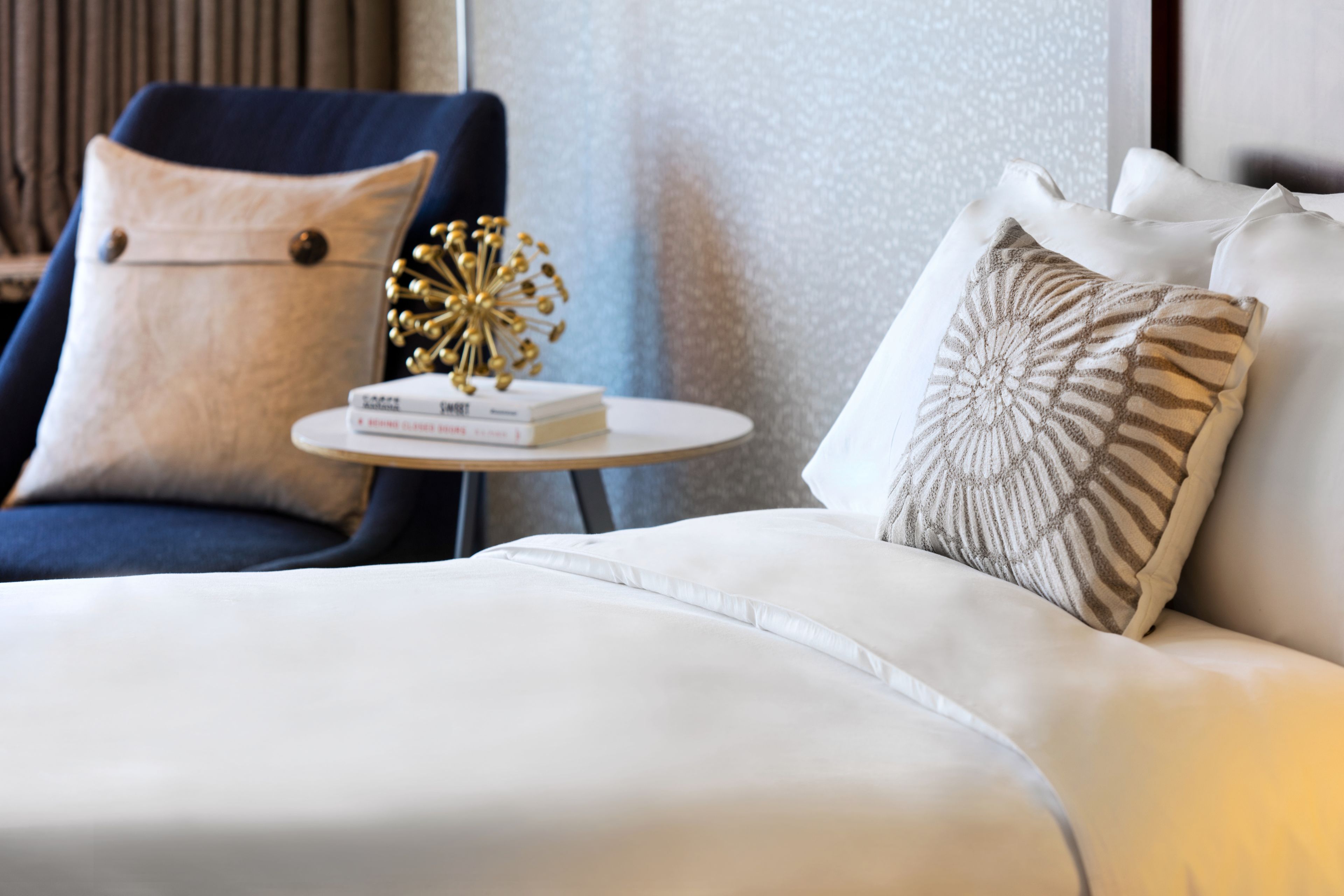 Una habitación de hotel moderna con un sofá blanco, un sillón azul y una mesita redonda con libros y un objeto decorativo.