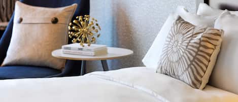 Una habitación de hotel moderna con un sofá blanco, un sillón azul y una mesita redonda con libros y un objeto decorativo.