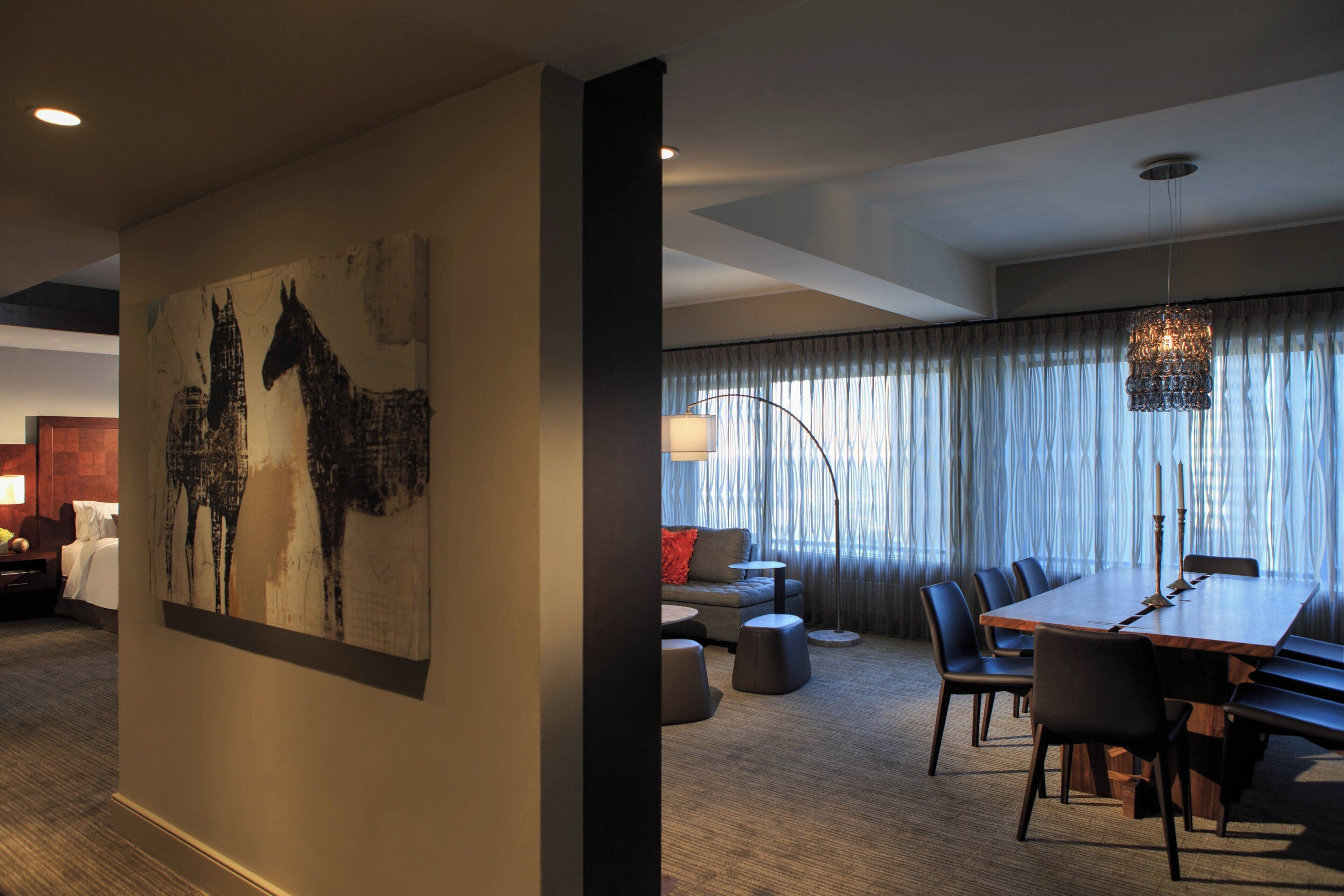 A modern hotel room with a large dining table, comfortable seating, a large abstract painting, and a view of the city through sheer curtains.