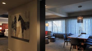 Una camera d'hotel moderna con un grande tavolo da pranzo, posti a sedere confortevoli, un'ampia pittura astratta e vista sulla città attraverso tende leggere.