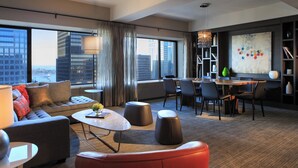 A modern hotel room with a large window, a sofa, a dining area, and a bookshelf.