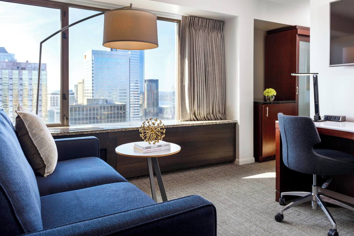 Ein modernes Hotelzimmer mit einem blauen Sofa, einem kleinen runden Tisch mit Büchern, einem Schreibtisch mit Stuhl, einer Lampe, einem Fenster mit Vorhängen und Blick auf Stadtgebäude.