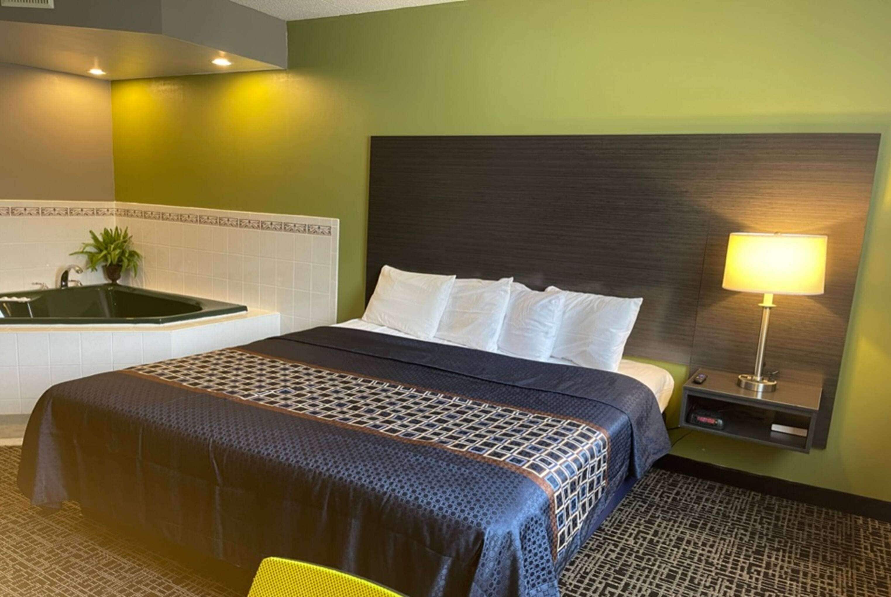 Quarto de hotel com cama, mesa de cabeceira com luminária, pequena mesa e televisão na parede.