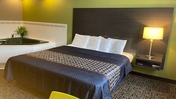 Quarto de hotel com cama, mesa de cabeceira com luminária, pequena mesa e televisão na parede.