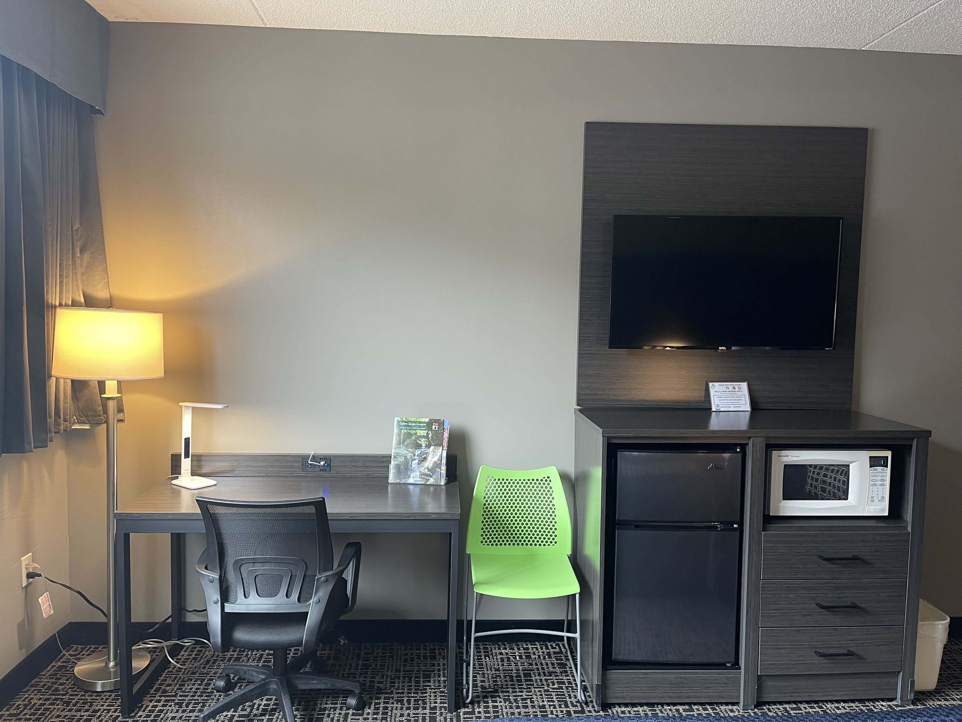 Habitación de hotel con escritorio, silla, lámpara, televisor, microondas y refrigerador pequeño.