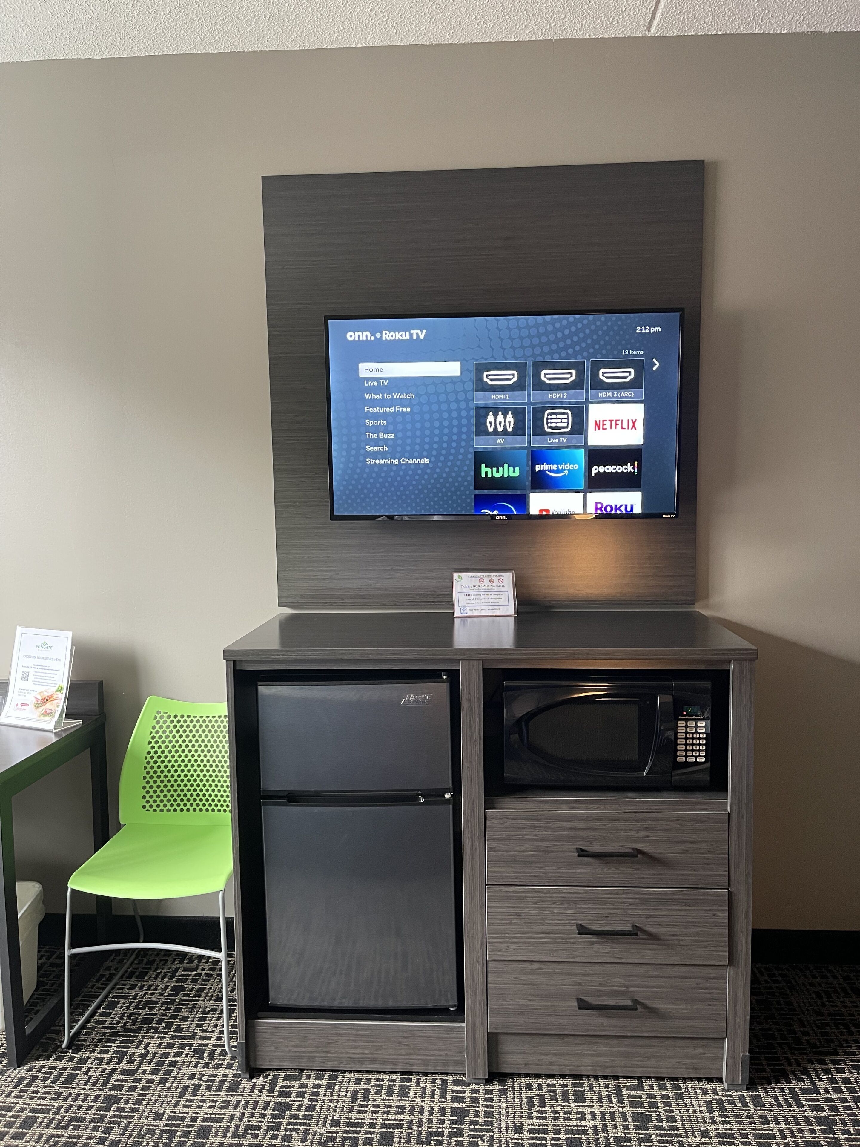 A hotel room with a built-in TV displaying a Roku interface, a mini-fridge, a microwave, and a green chair.