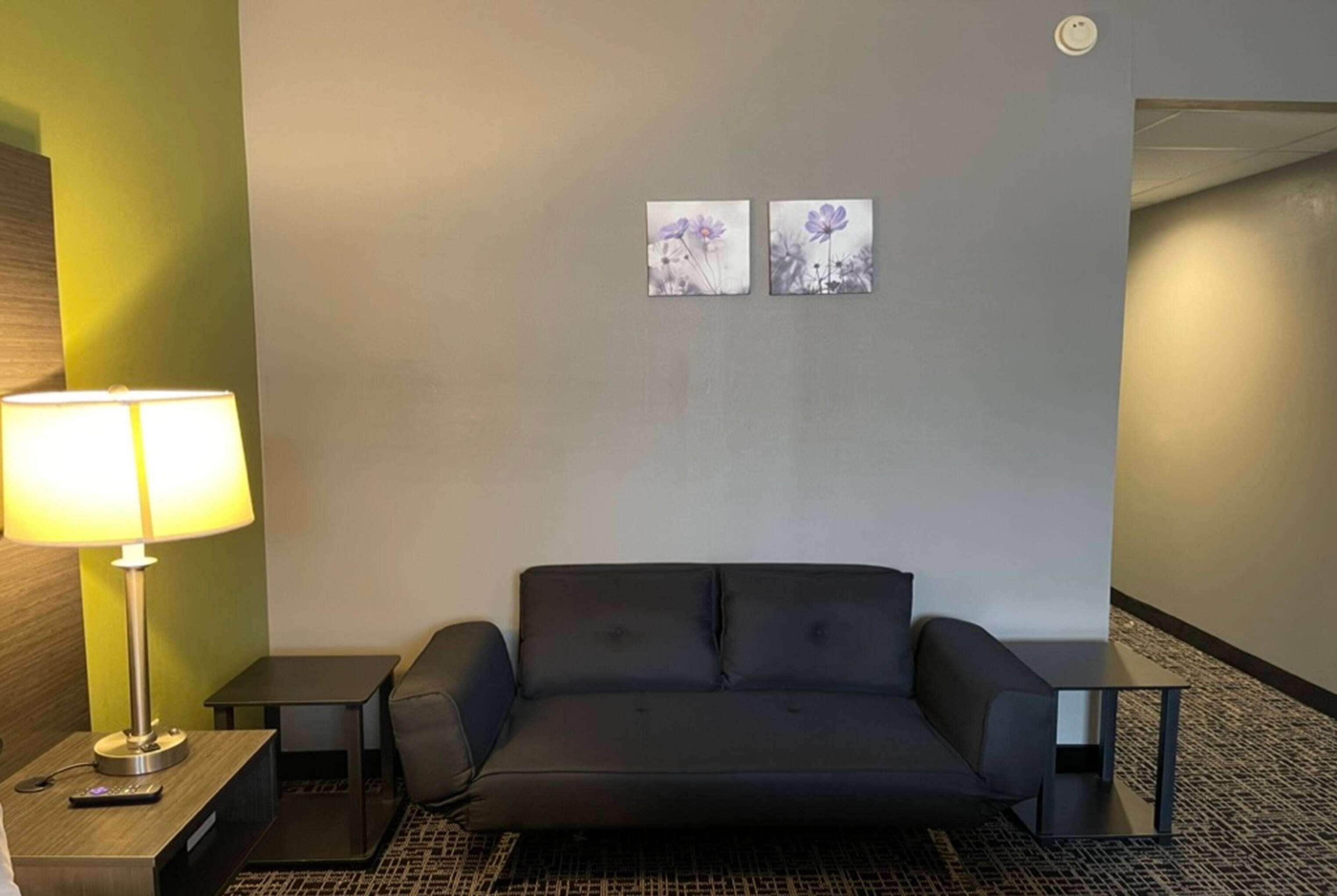 A hotel room with a black sofa, a lamp, two small tables, and two wall-mounted flower paintings.