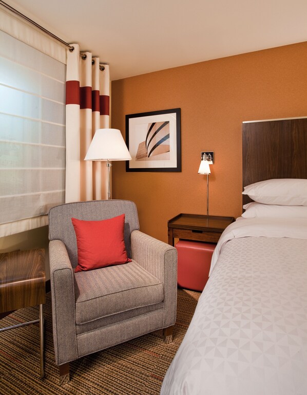 A hotel room with a bed, a chair with a red pillow, a bedside table with a lamp, and a framed picture on the wall. - The Crossroads Hotel, Newburgh (Newburgh)