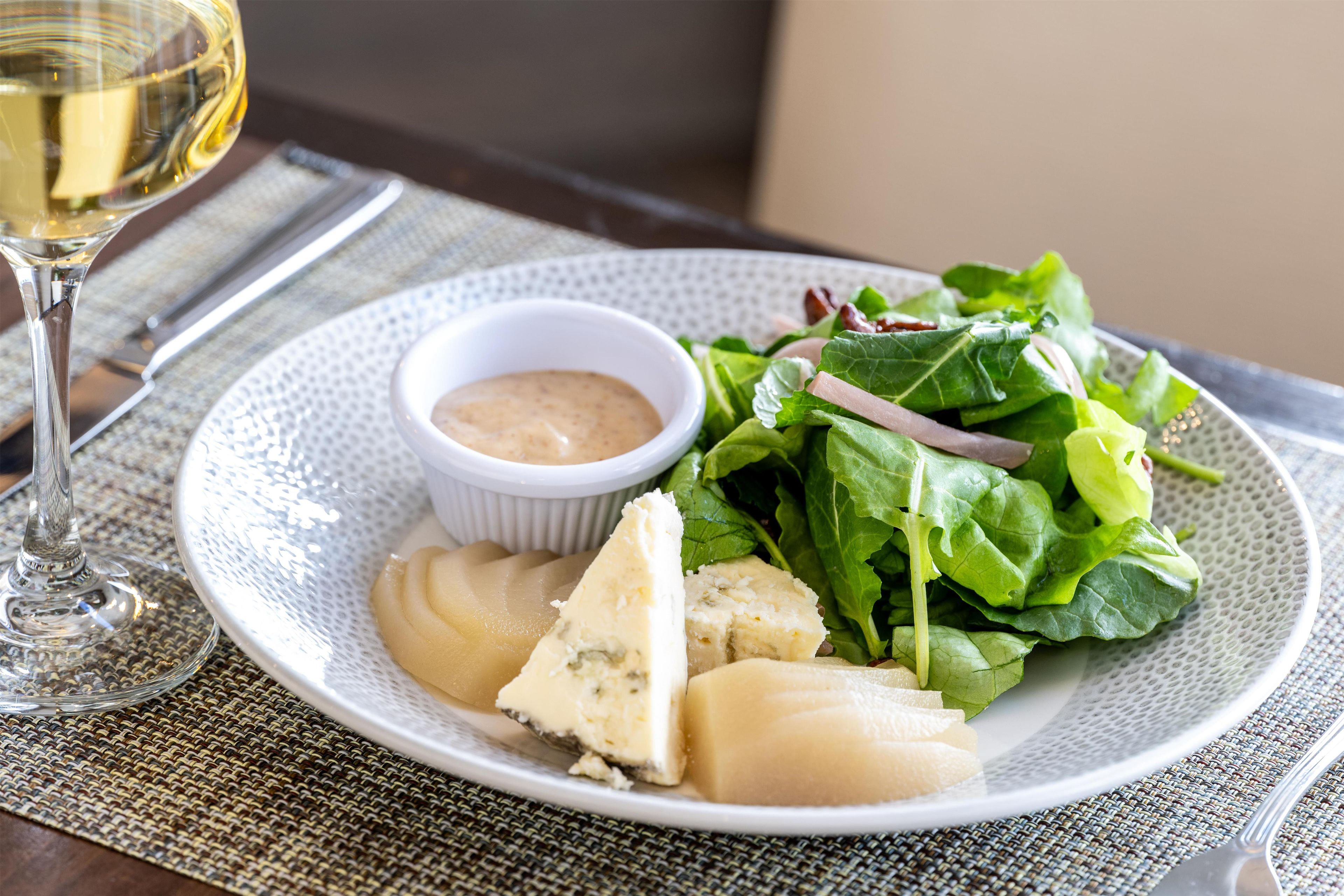 Un plato con ensalada, queso y un recipiente pequeño de salsa, acompañado de una copa de vino.