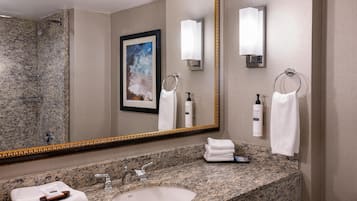 A bathroom with a granite countertop, a sink, a mirror with a gold frame, a framed painting, and two towel rings.