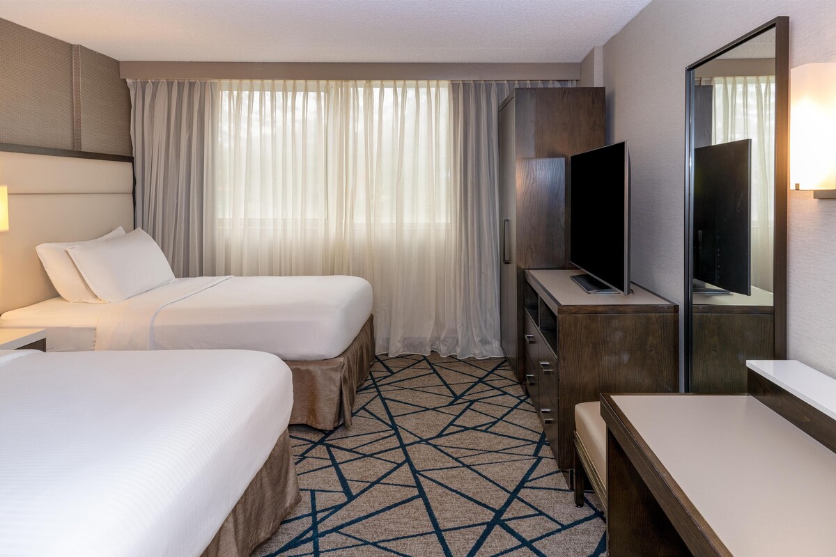 A hotel room with two beds, a TV, a desk, and a large window with curtains.