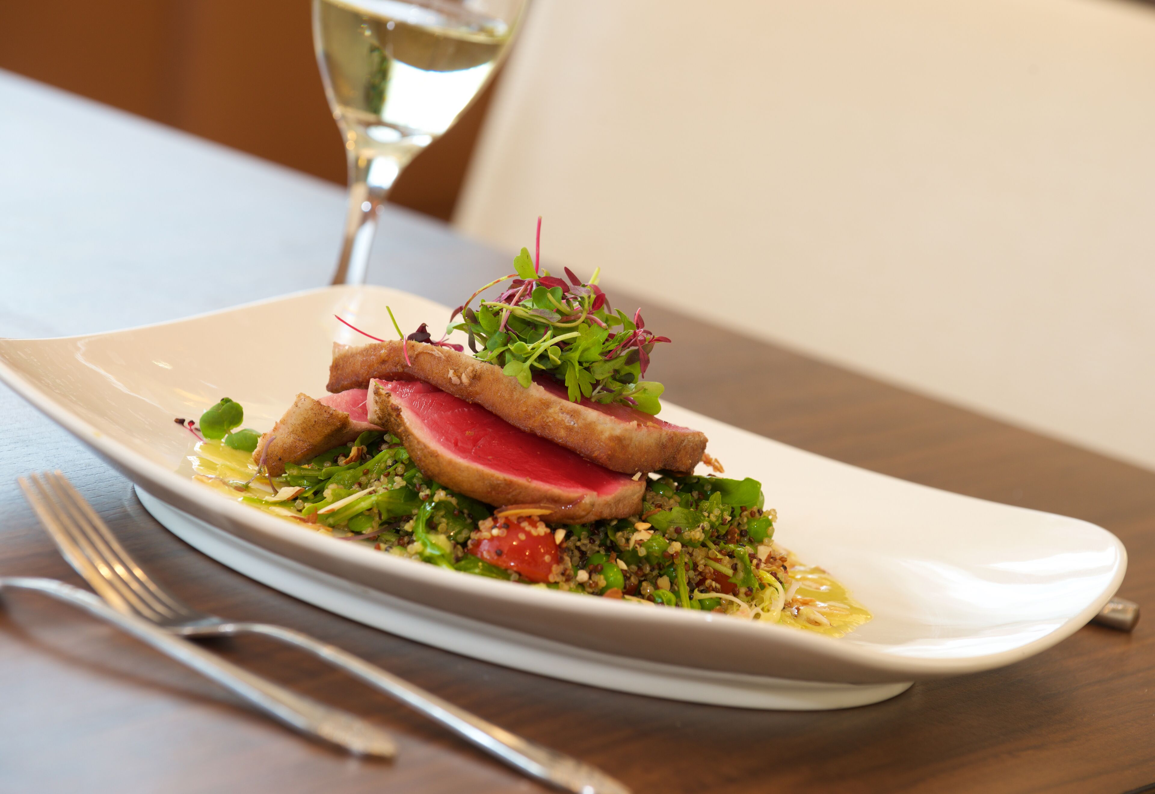 A white plate with a dish of meat, greens, and a small salad, accompanied by a glass of white wine.