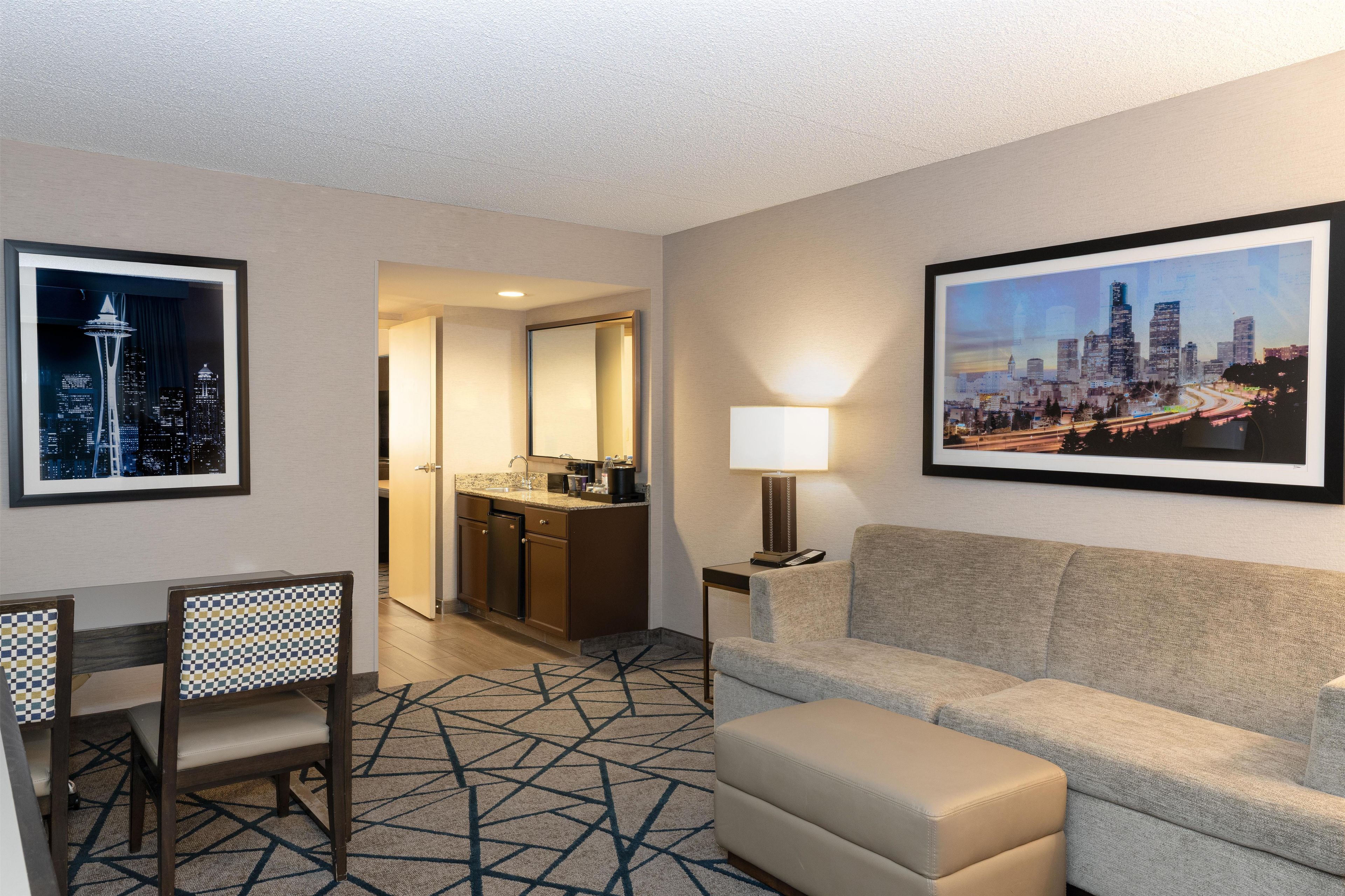 A hotel room with a sofa, a dining table with chairs, a kitchen area, and two framed cityscape pictures on the walls.