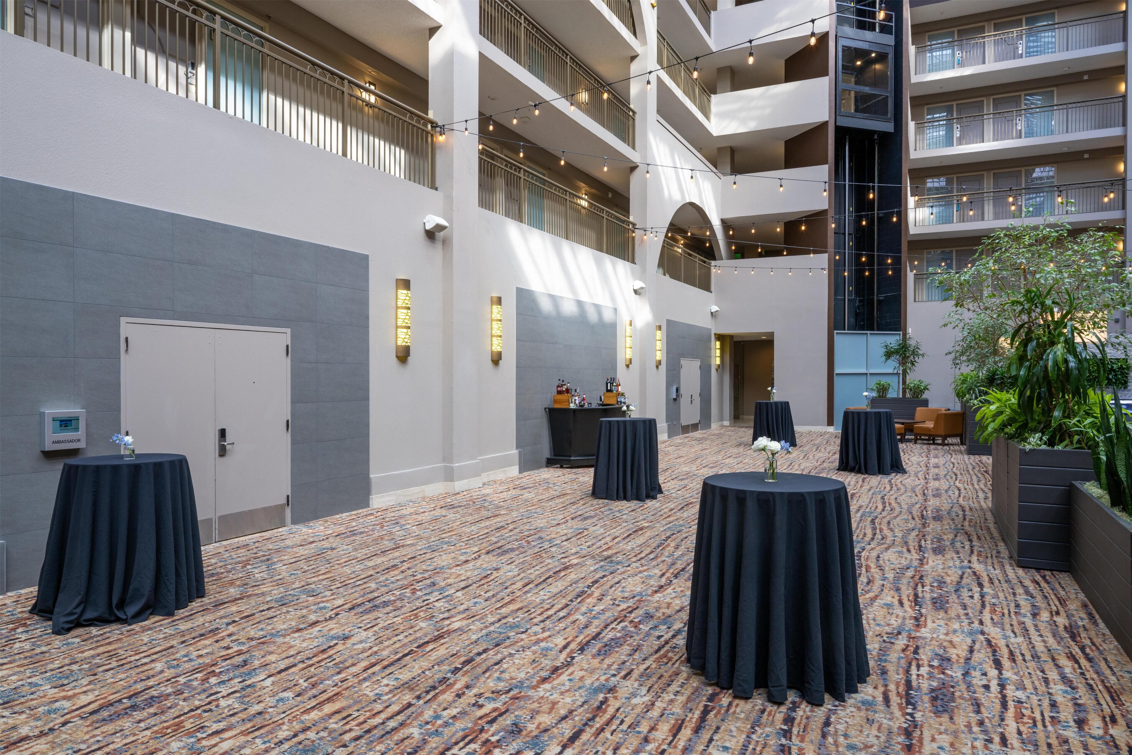A spacious indoor area with multiple levels, featuring a patterned carpet, tables with dark coverings, and a balcony with string lights.
