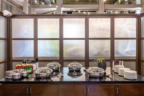 A buffet setup with food containers, plates, and utensils on a counter with a large window in the background. - Embassy Suites by Hilton Seattle Bellevue (Bellevue)