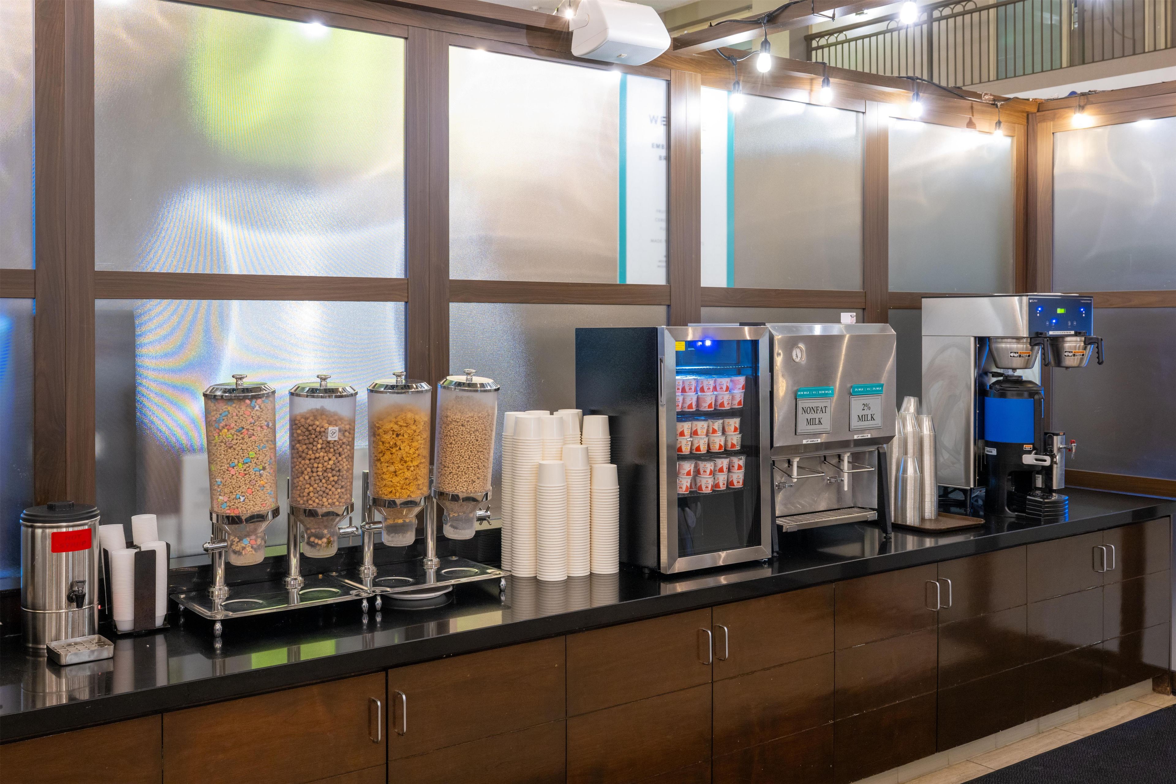 A self-service breakfast bar with dispensers for cereal, milk, and coffee, and a refrigerated display case.