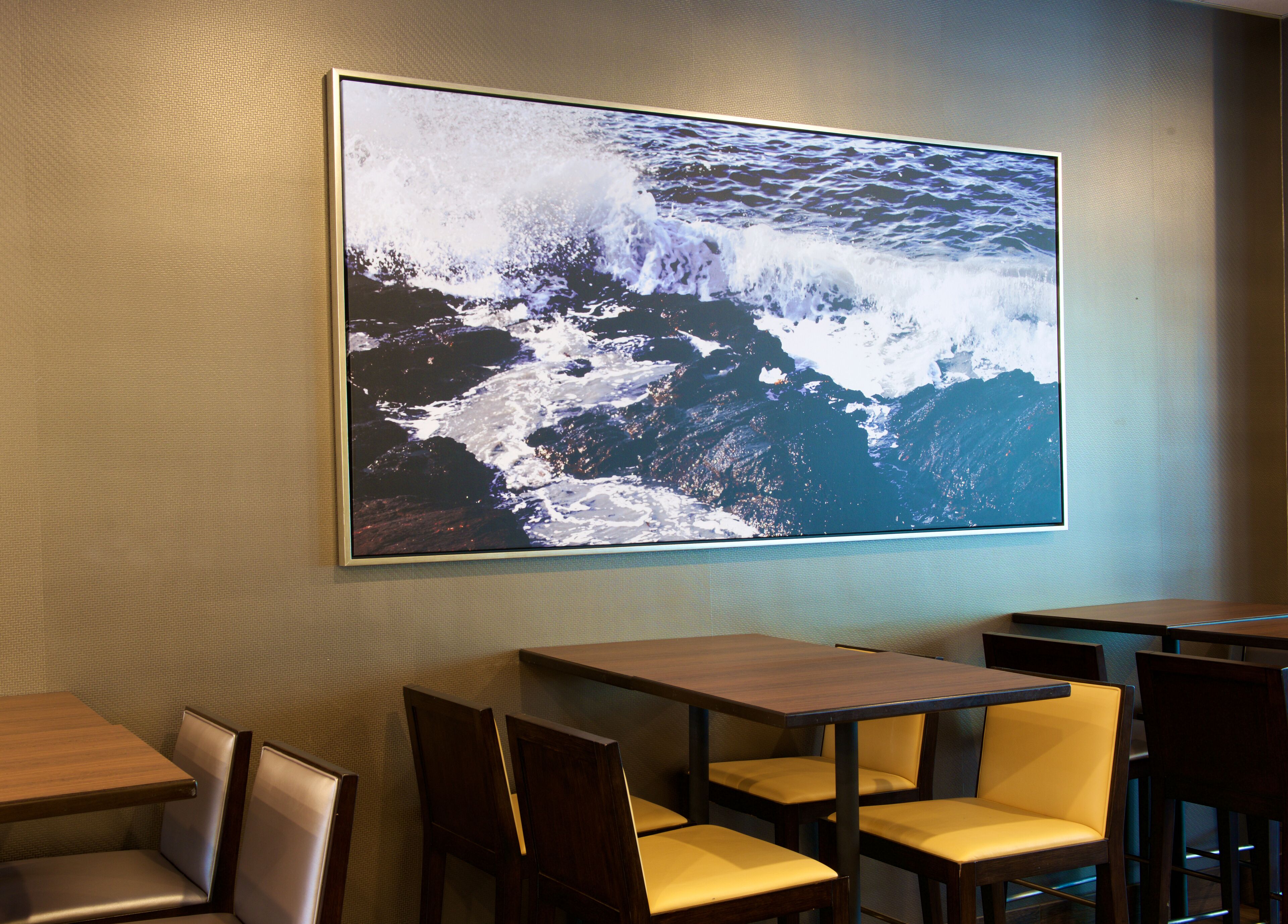 A restaurant with a large ocean wave painting on the wall, wooden tables, and yellow cushioned chairs.