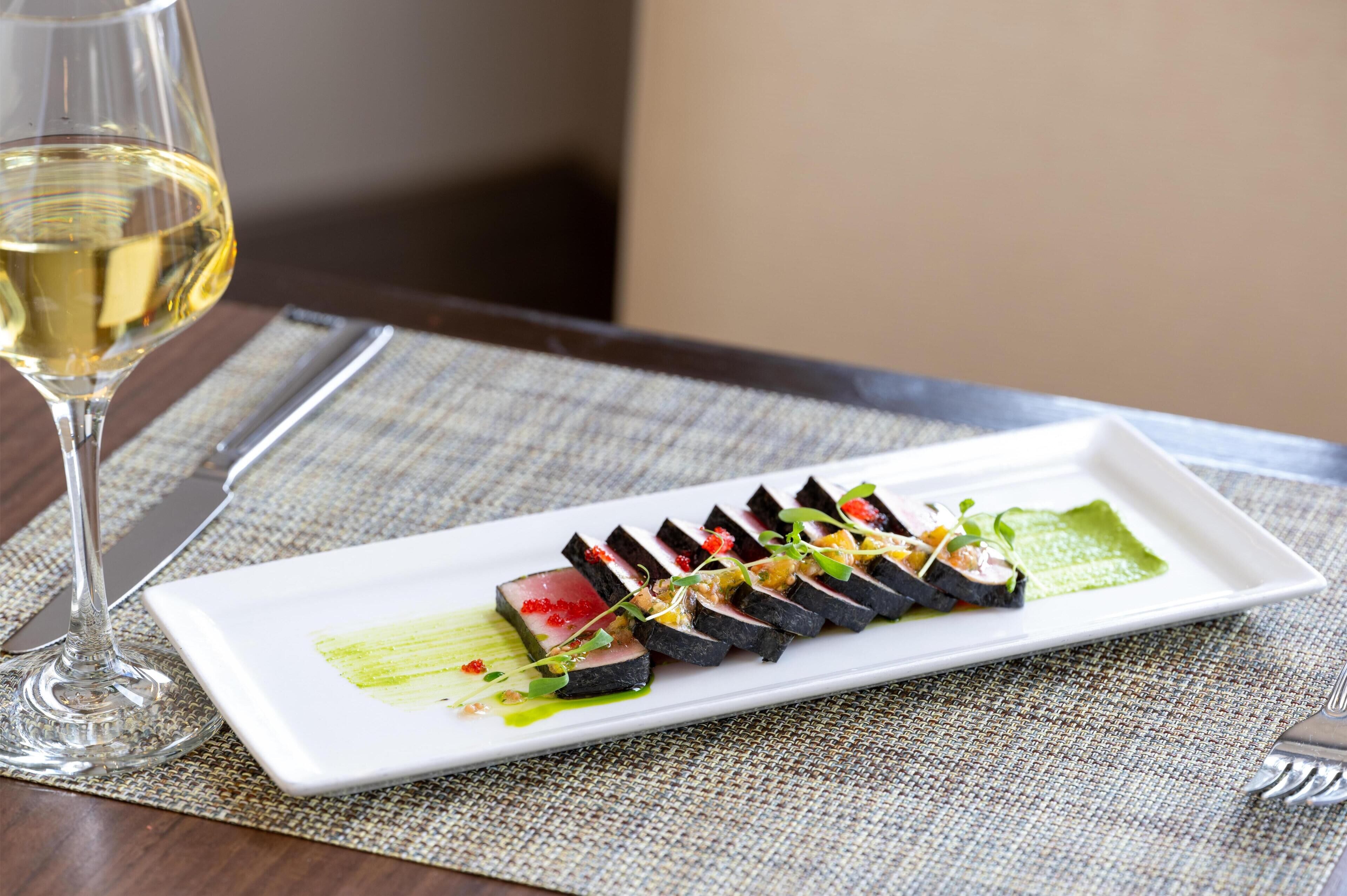 A rectangular white plate with a sushi dish, a glass of white wine, and a fork on a textured tablecloth.