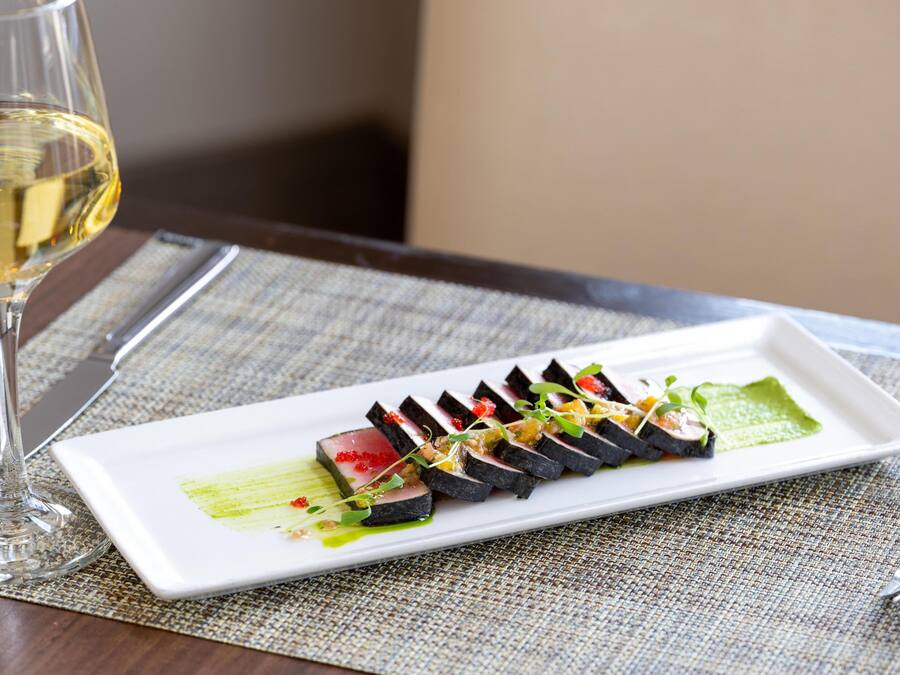 A rectangular white plate with a sushi dish, a glass of white wine, and a fork on a textured tablecloth.