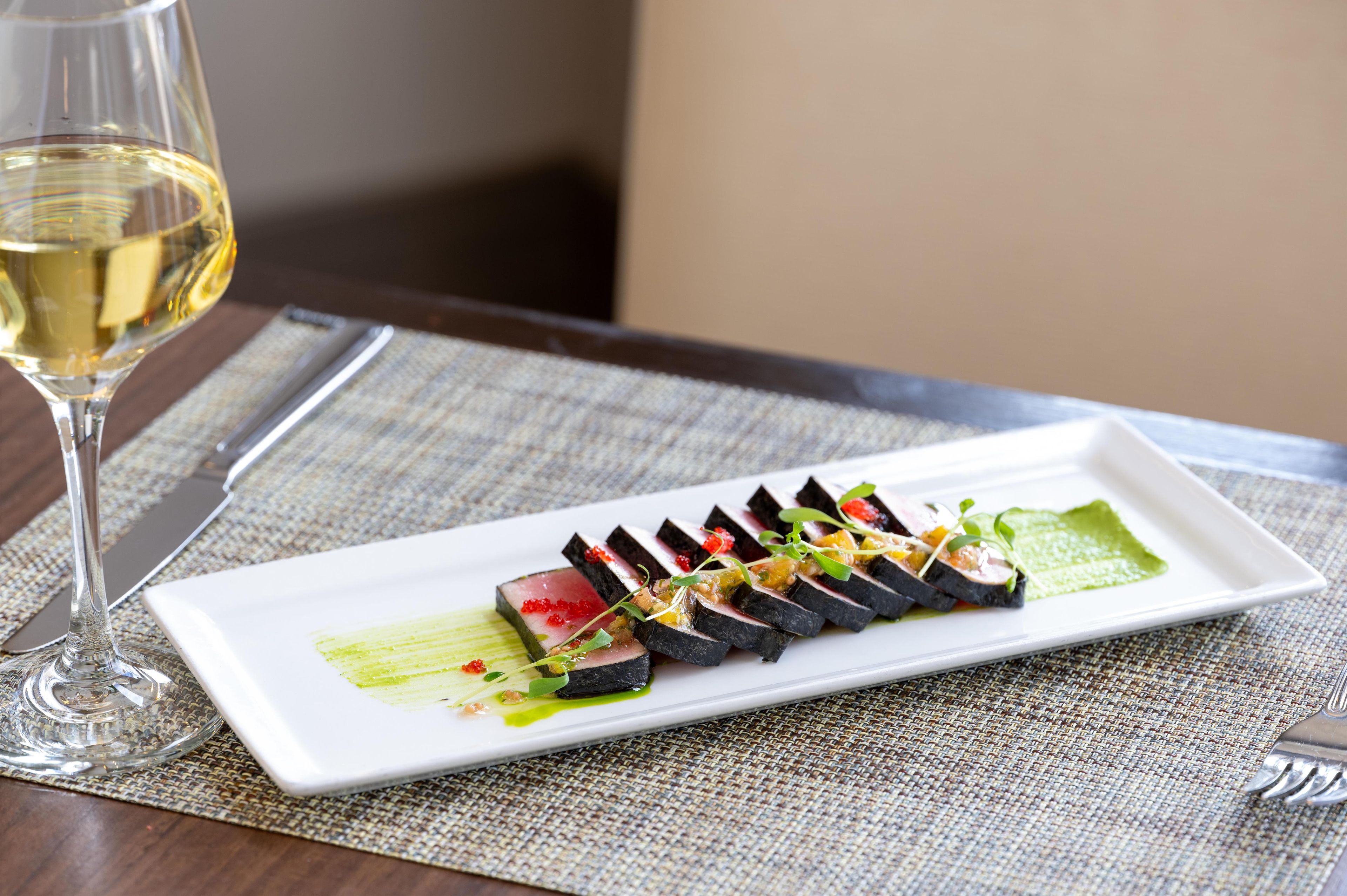 A rectangular white plate with a sushi dish, a glass of white wine, and a fork on a textured tablecloth.