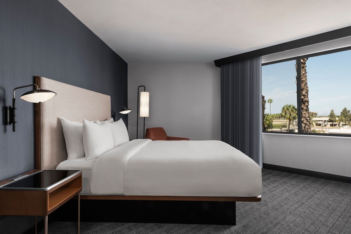 Une chambre d’hôtel moderne avec un grand lit, deux tables de chevet, une petite table et une vue sur les palmiers, visible à travers une grande fenêtre.