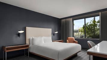 A modern hotel room with a large bed, two bedside tables, a desk, and a chair. There are two wall-mounted lamps and a window with curtains.