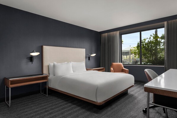 A modern hotel room with a large bed, two bedside tables, a desk, and a chair. There are two wall-mounted lamps and a window with curtains.