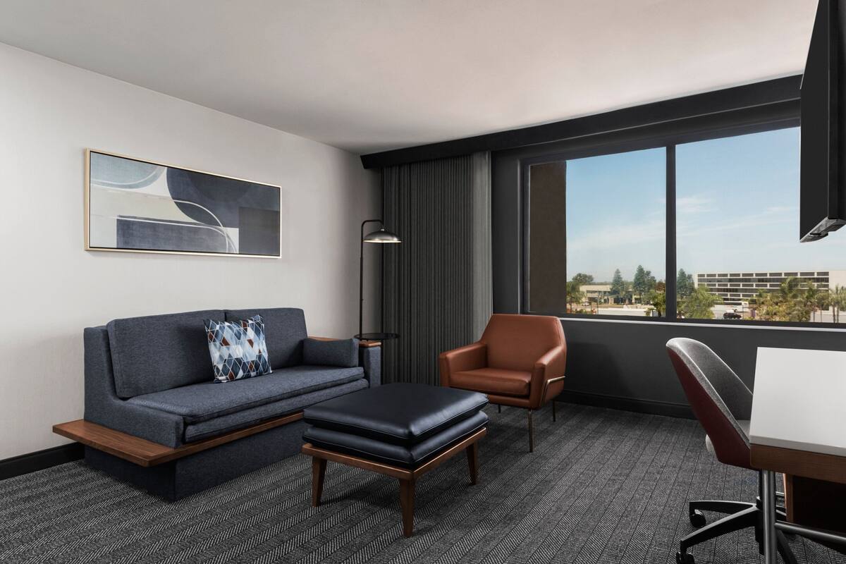 Une chambre d’hôtel moderne avec un canapé gris, un fauteuil en cuir marron, un pouf noir, un bureau et une grande fenêtre donnant sur la ville.