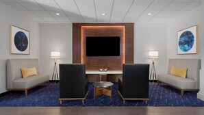 A hotel lobby with a flat-screen TV mounted on a wooden panel, two sofas with yellow cushions, a small round table, and two floor lamps. - Courtyard by Marriott San Diego Miramar (San Diego)