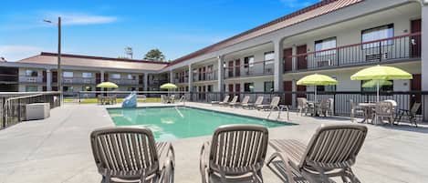 Un área de piscina con sillones y sombrillas, un edificio con varios balcones y un cielo despejado.
