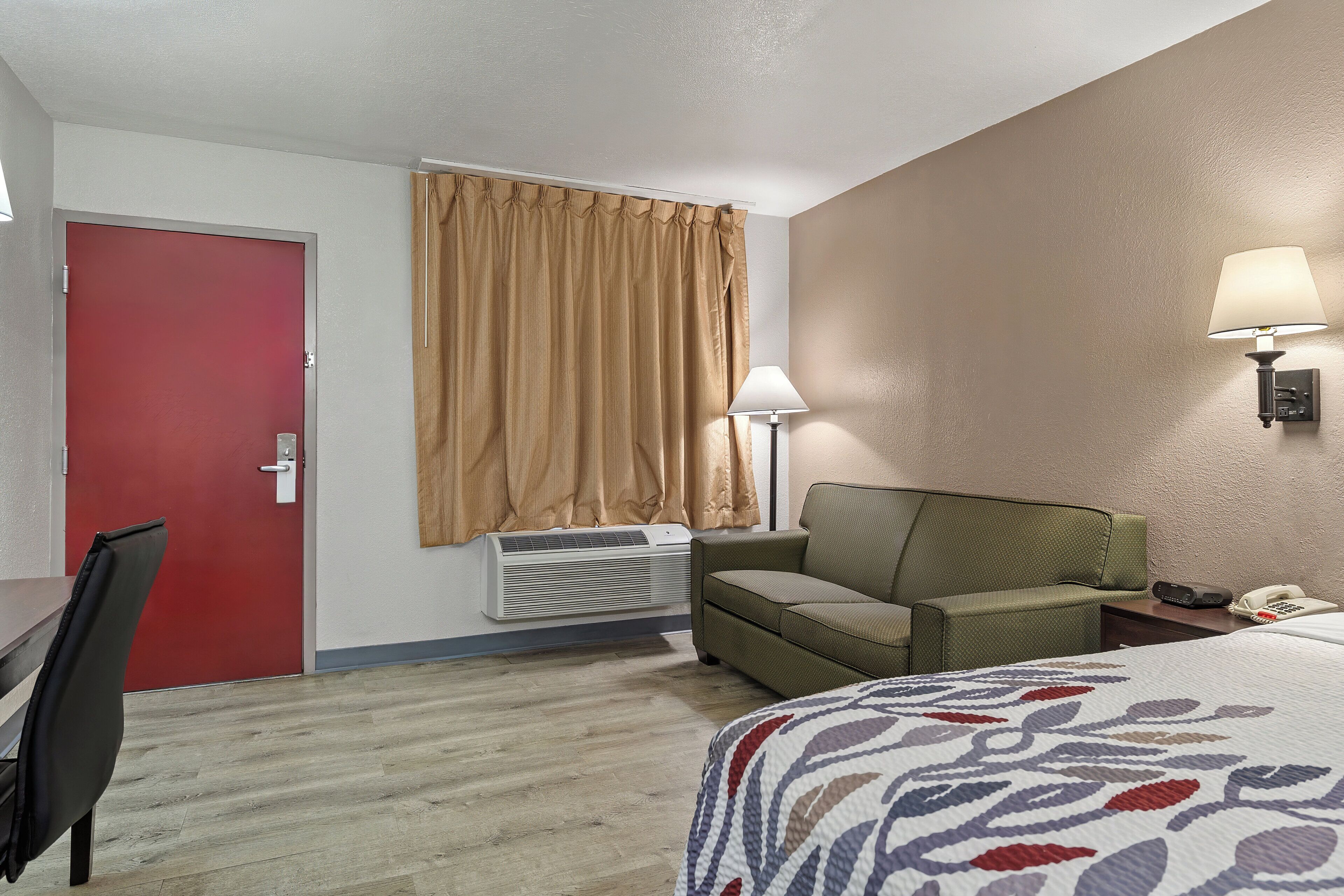 Une chambre d’hôtel avec un lit, une chaise, un bureau, une porte rouge, un climatiseur, une lampe et un téléphone.