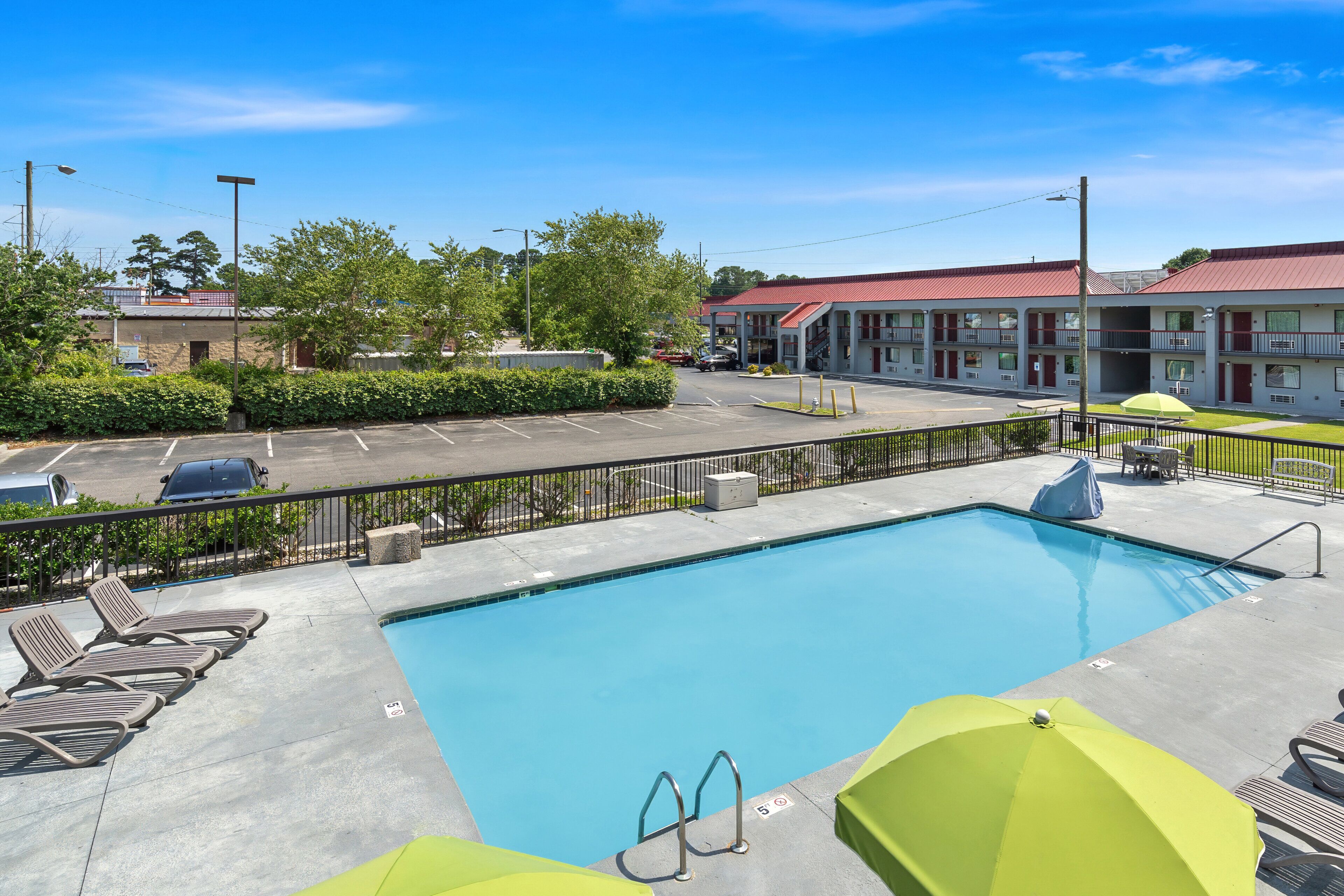 Ein Swimmingpool mit Liegestühlen und Sonnenschirmen, umgeben von einem eingezäunten Parkplatz und einem Motelgebäude.