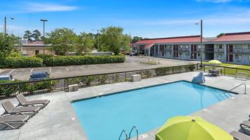 A swimming pool with lounge chairs and umbrellas, surrounded by a fenced parking area and a motel building.