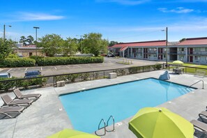 Una piscina con sillones de descanso y sombrillas, rodeada de un estacionamiento cercado y un edificio de motel.
