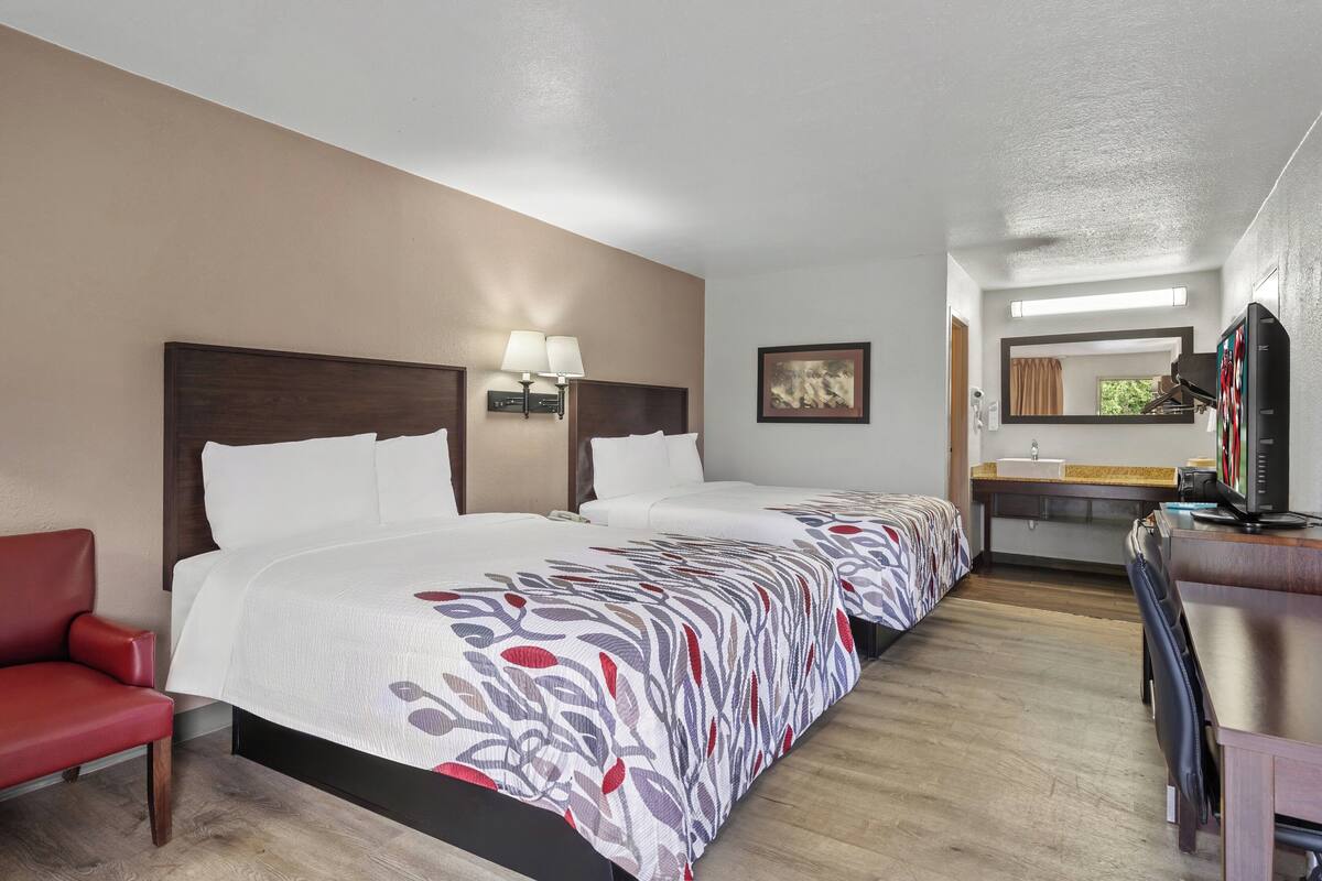 Ein Hotelzimmer mit zwei Betten, einem Schreibtisch, einem Sessel, einem Fernseher und einem im Hintergrund sichtbaren Badezimmer.