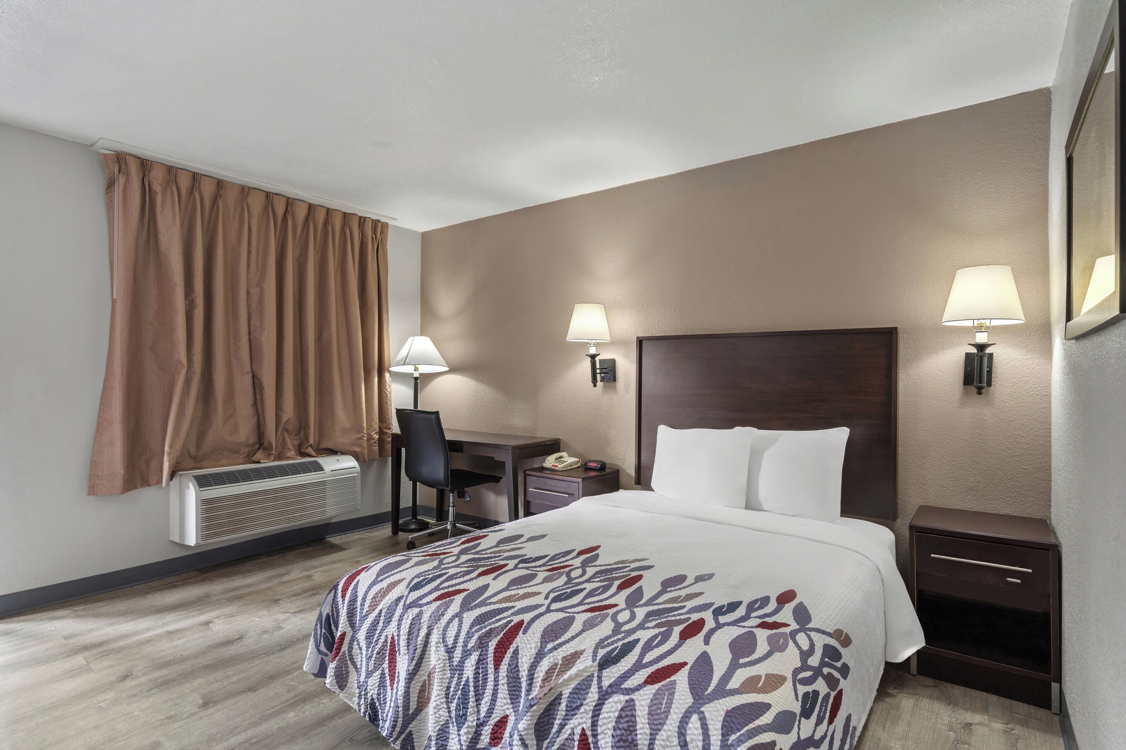 A hotel room with a bed, desk, chair, bedside lamps, and a window with curtains.