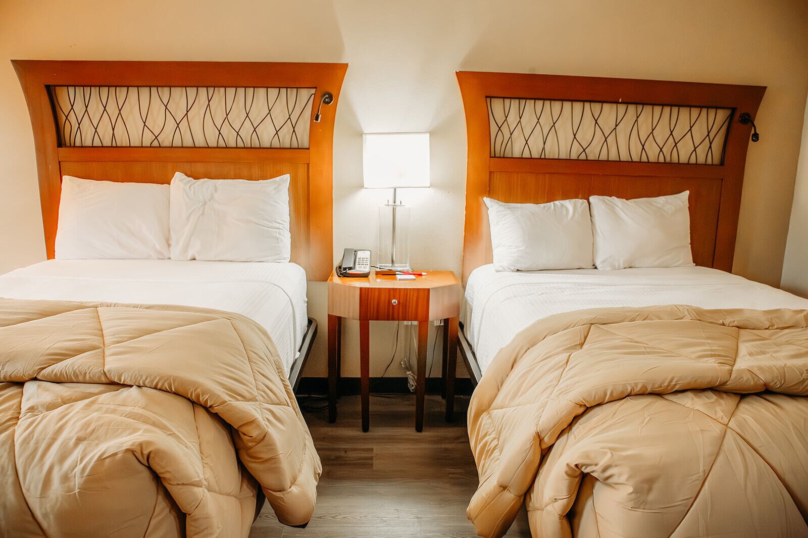 Een hotelkamer met twee bedden, elk met een houten hoofdbord en witte beddengoed. Er staat een klein nachtkastje met een lamp en een telefoon.