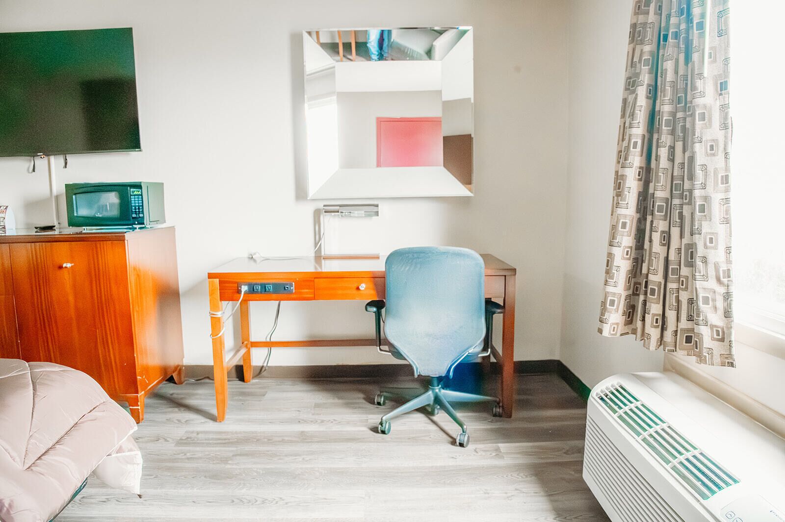 A room with a wooden desk, a blue office chair, a television on the wall, a radiator, and a window with patterned curtains.